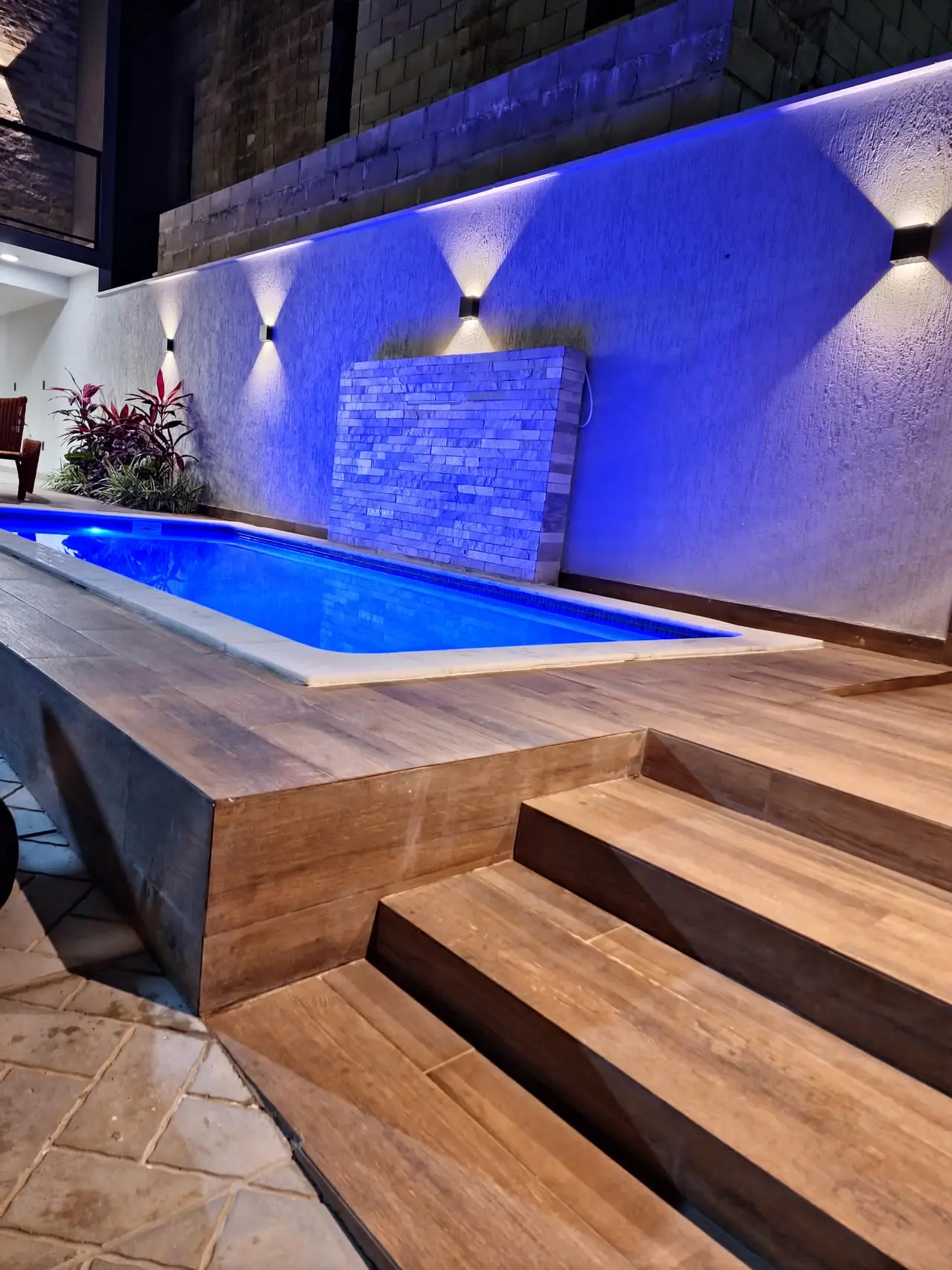 Piscina iluminada à noite, com cascata de pedra ao fundo e deck de madeira