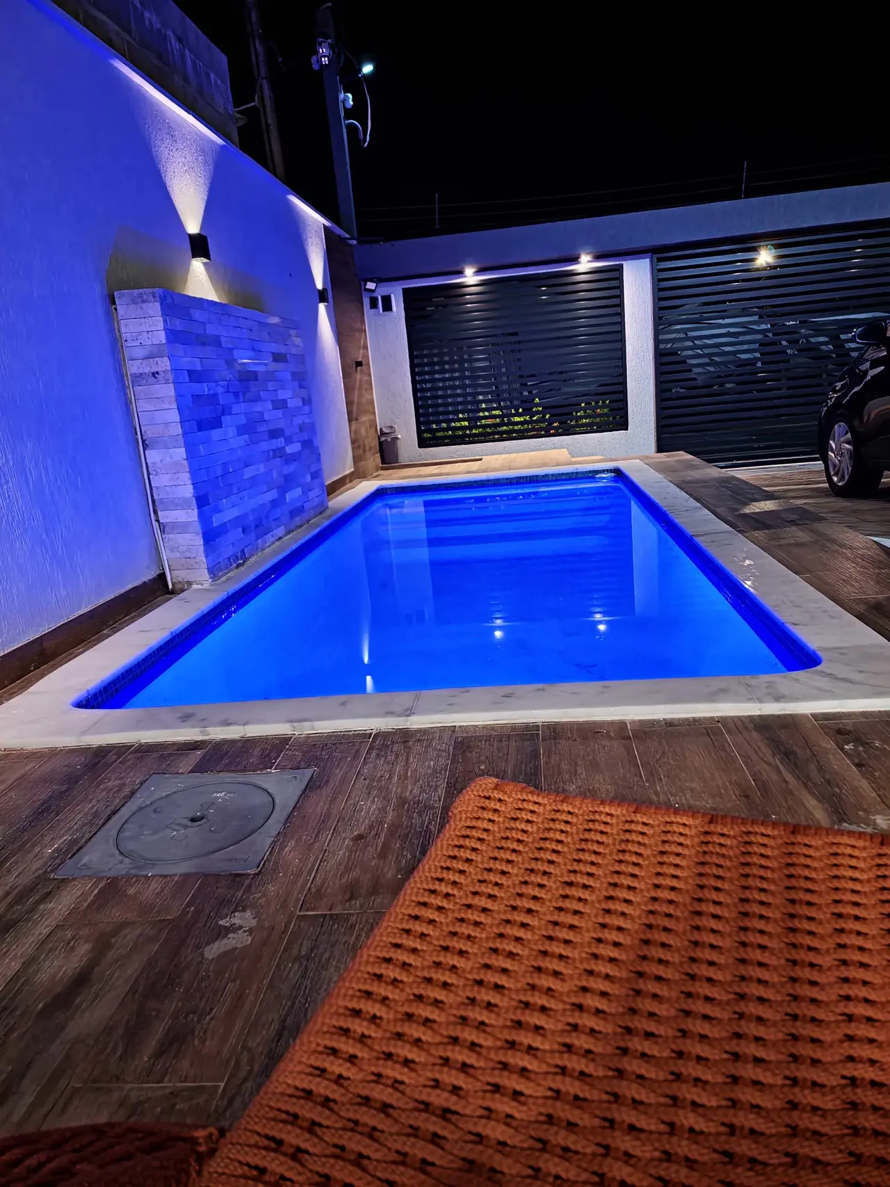 Piscina iluminada em azul à noite, com a cascata e o deck de madeira