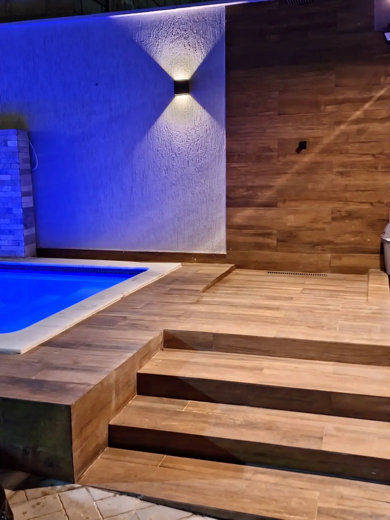 Deck de madeira com degraus ao lado da piscina, sob luz de parede